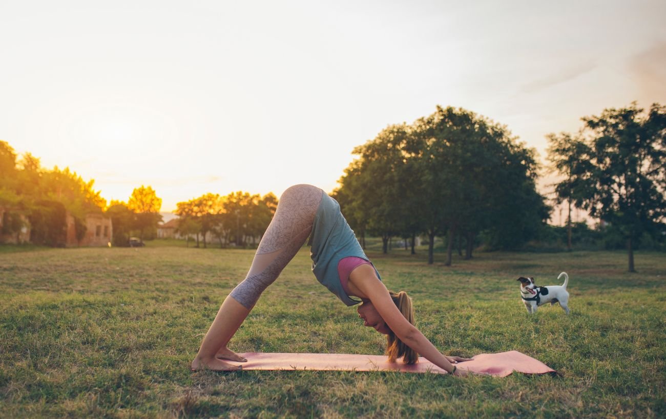 a woman doing downward dog in a park with a dog next to her