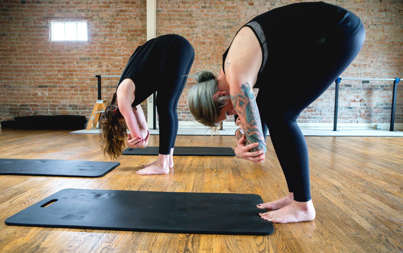 two women doing a standing forward fold in a yoga studio