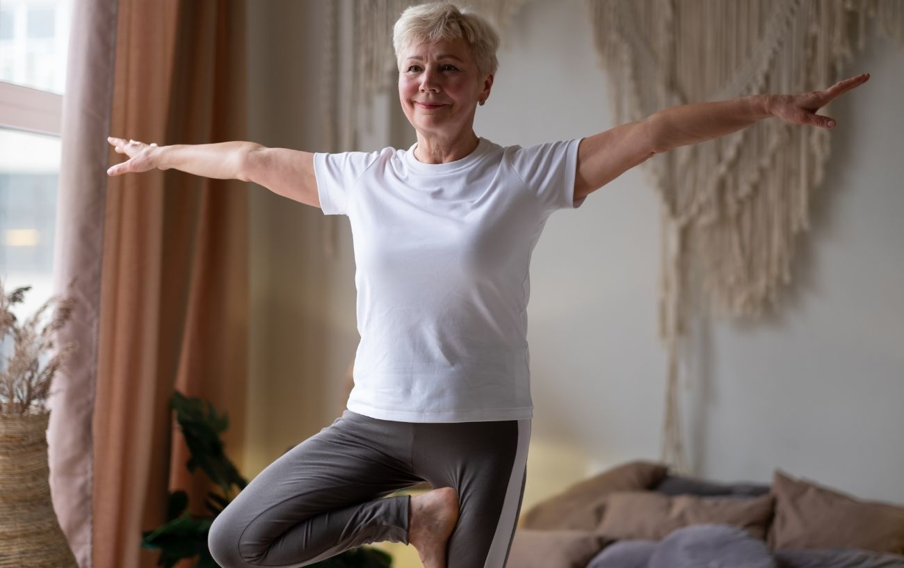 an elderly woman doing tree pose in her bedroom in a white top