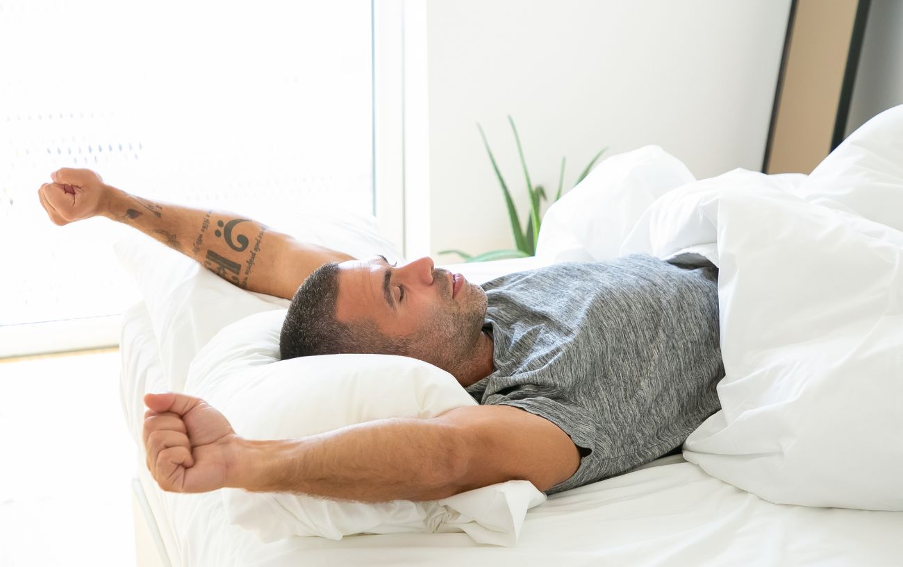 A man in a grey t shirt stretching in a white bed.