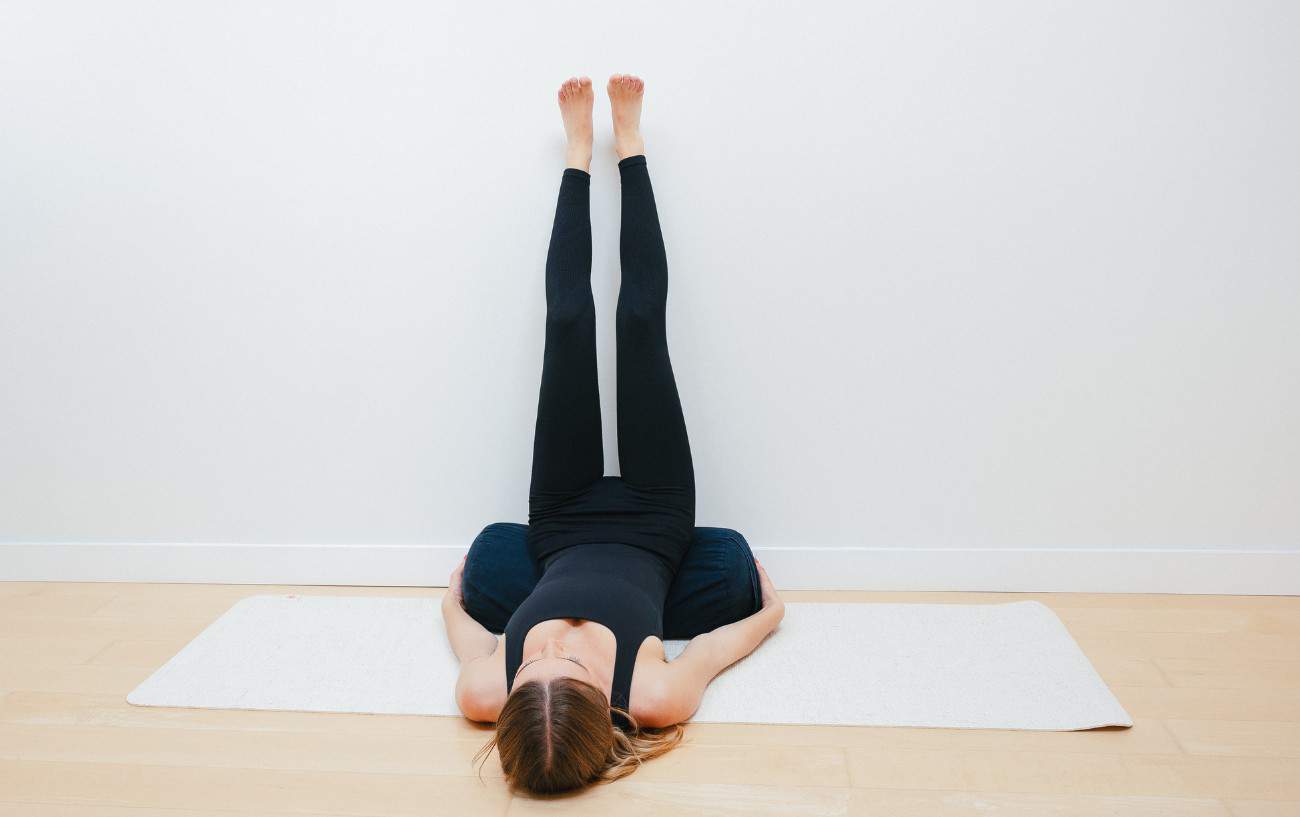 woman doing legs up the wall yoga pose with a bolster under her hips in a yoga studio