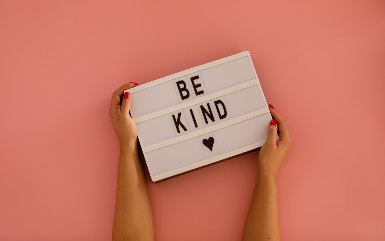 Hands holding a sign saying be kind against a pink background.