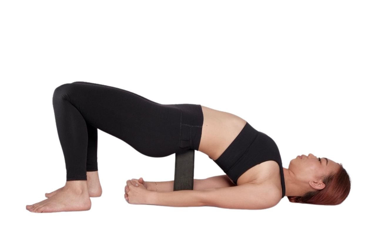 a woman wearing black yoga clothes in supported bridge pose with a block under her hips