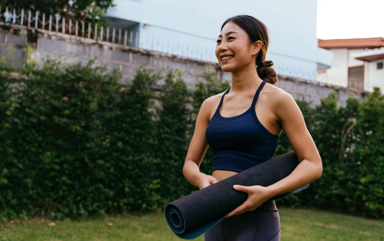 woman smiles and holds yoga mat in a garden